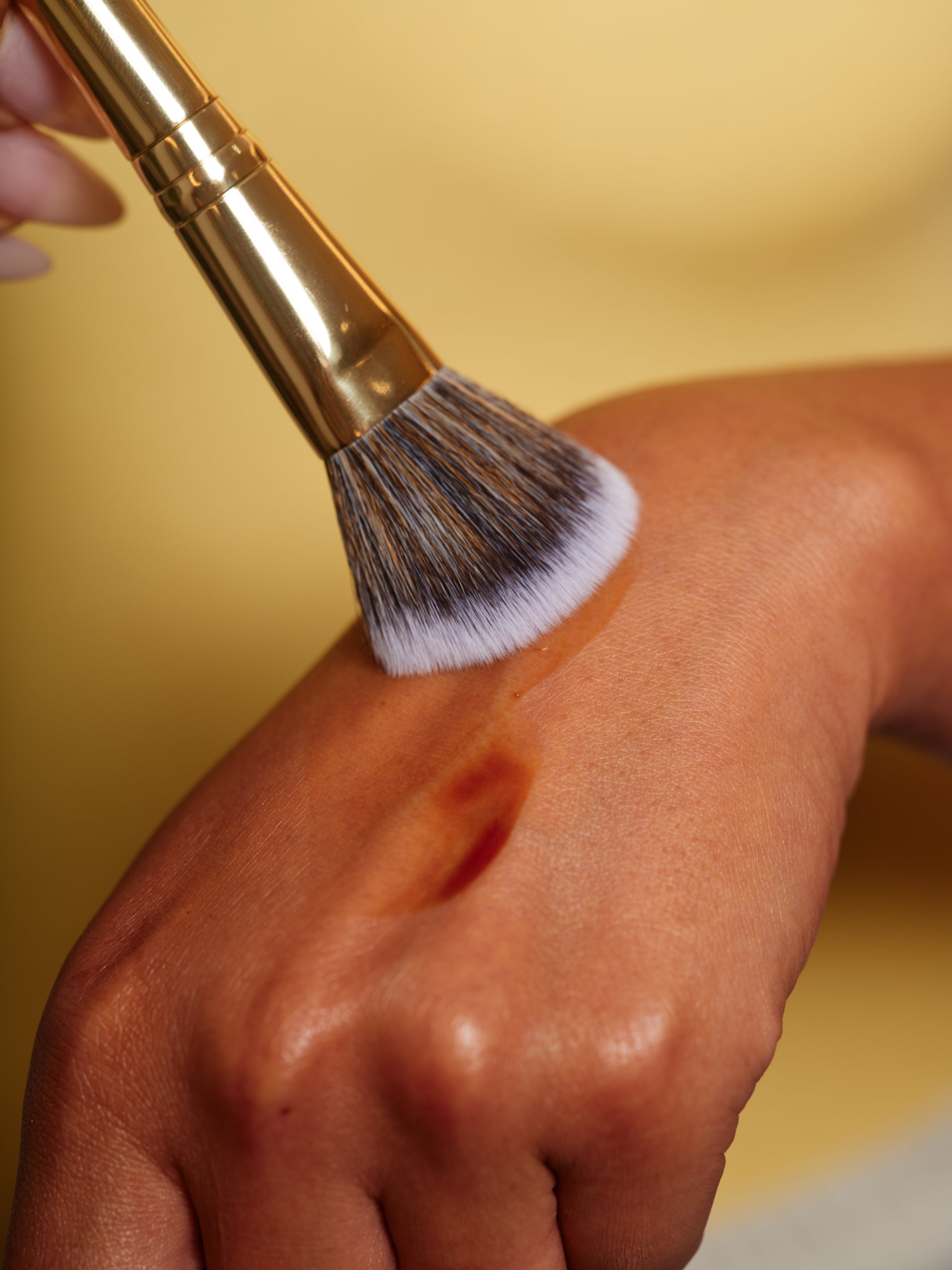 A makeup brush applies liquid foundation on the back of a persons hand against a yellow background.
