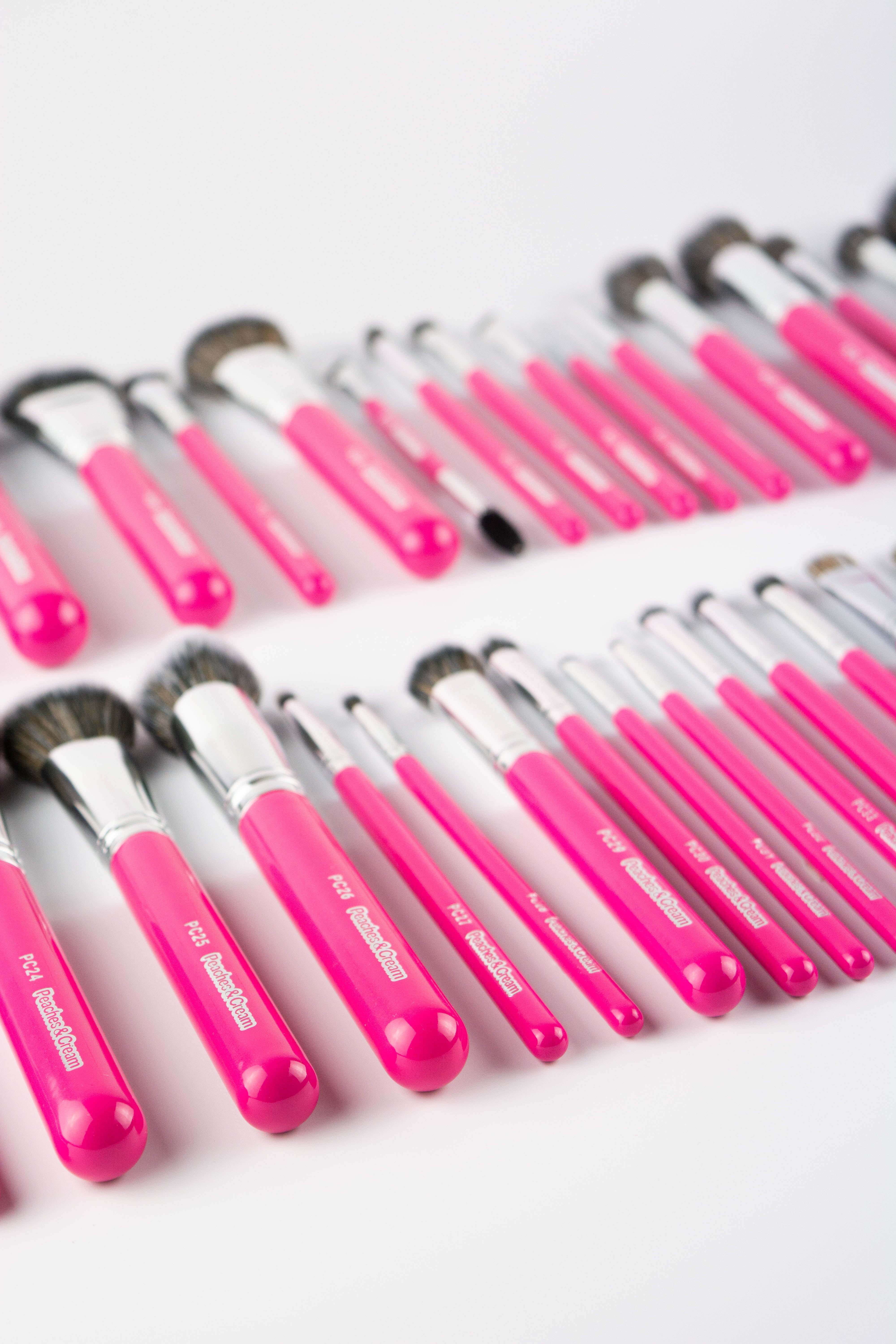 A collection of pink-handled makeup brushes of various sizes and shapes are arranged in a spiral pattern on a bright turquoise background.
