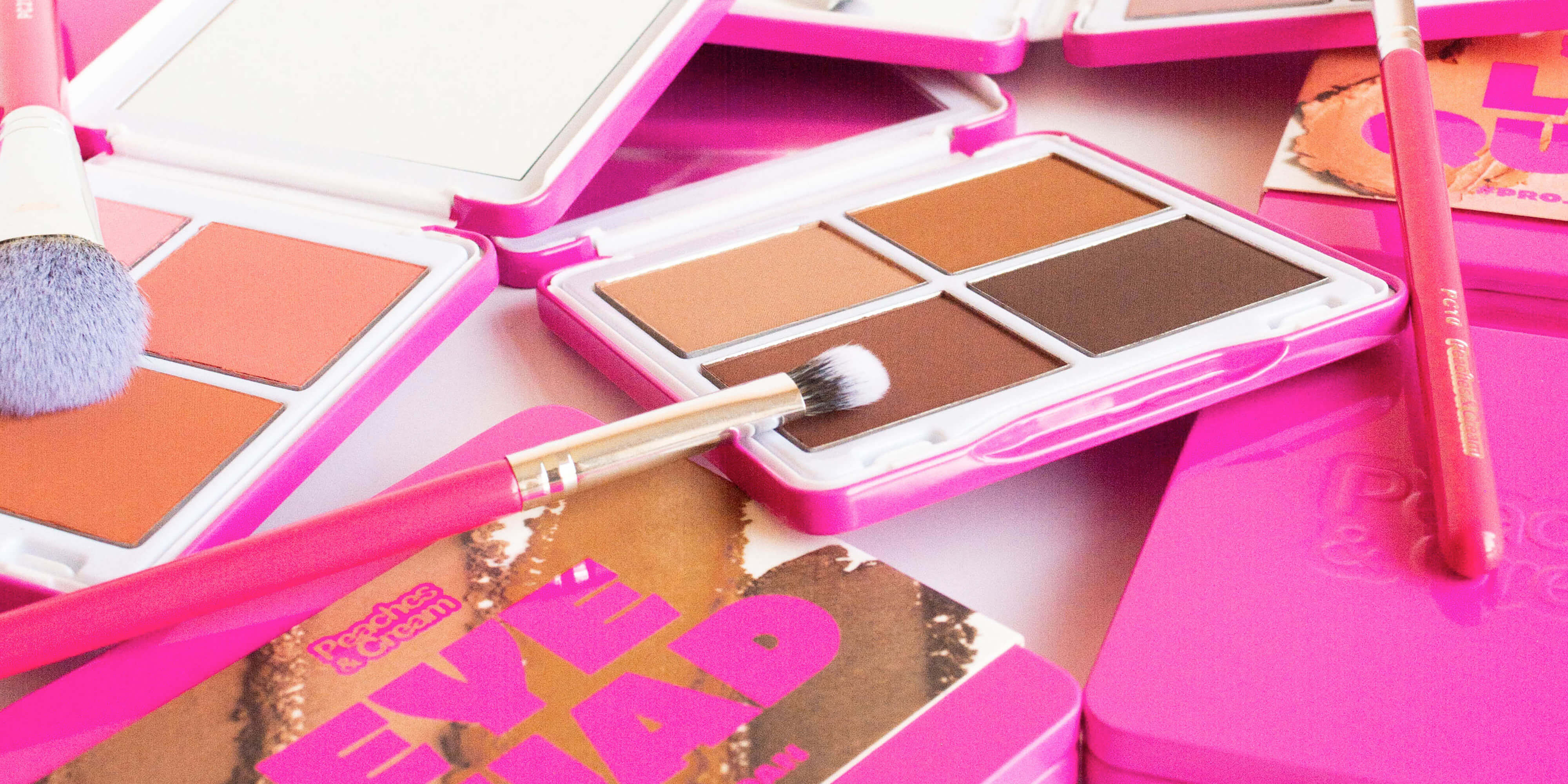 A close-up of brightly colored pink makeup palettes, including eyeshadows and blushes, with makeup brushes resting on and around the palettes.