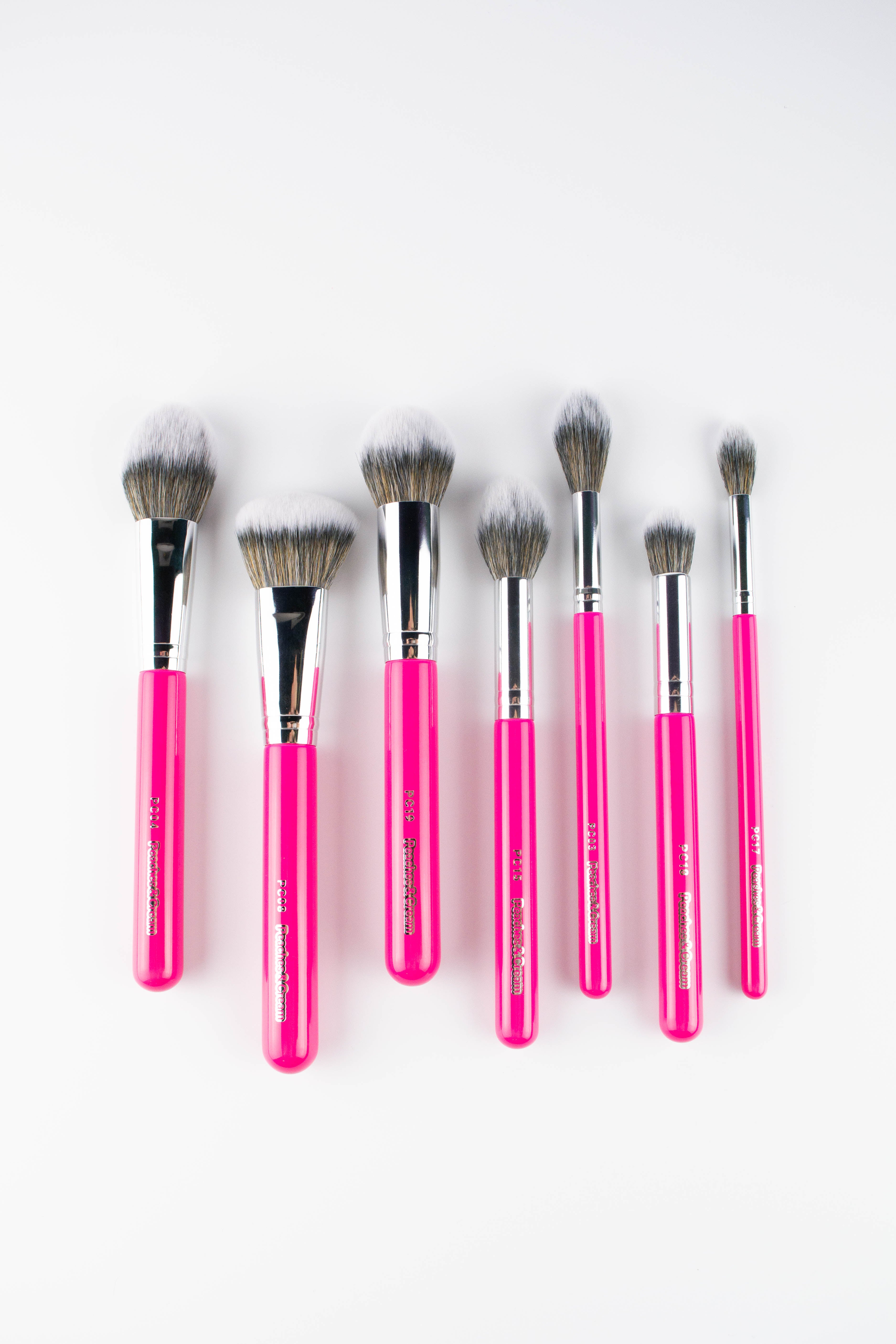 Six pink-handled Peaches and Cream Essential Face Brush Set brushes, featuring fluffy bristles, are arranged in a row on a white surface - ideal for flawless makeup with sizes and shapes for every step.