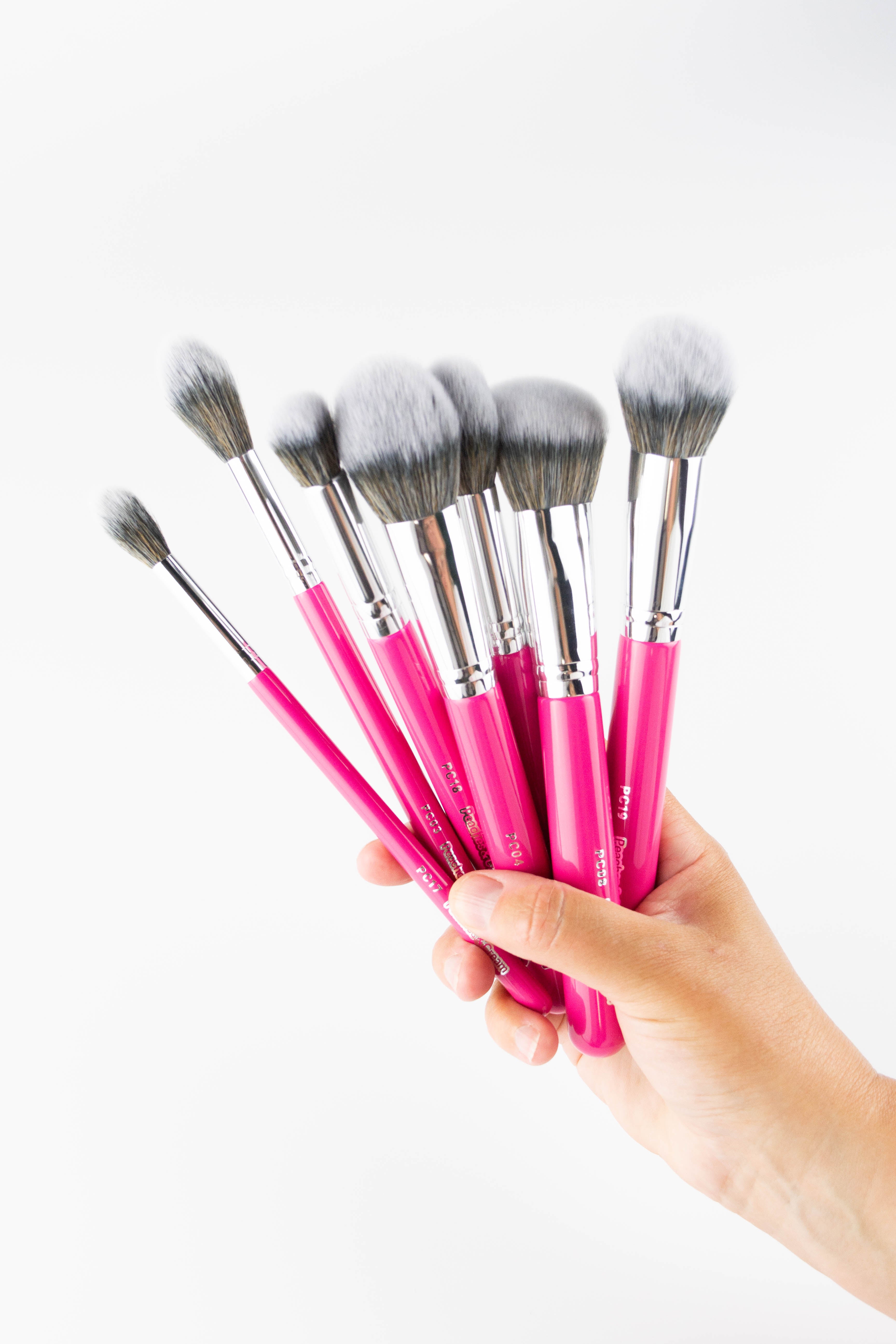 A hand holds the Peaches and Cream Essential Face Brush Set, featuring pink handles and soft white-gray bristles - perfect for flawless makeup - against a white background.