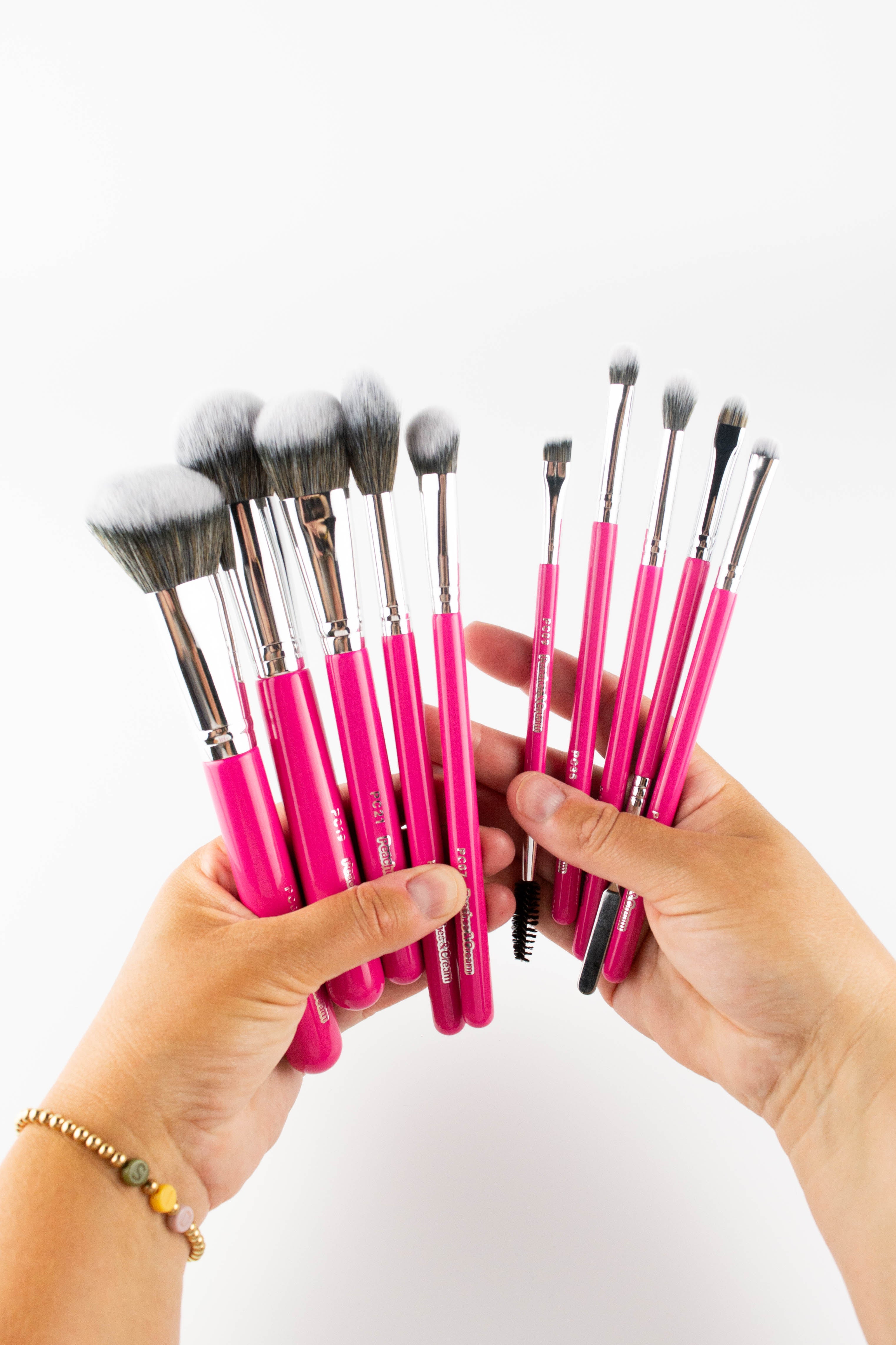 Two hands hold The Top 10 Makeup Brush Set by Peaches & Cream Makeup, featuring bright pink handles, silver ferrules, and soft synthetic fibres. The set includes varied brush sizes and shapes for different makeup applications against a white background.