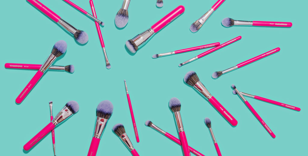 A collection of pink-handled makeup brushes of various sizes and shapes are arranged in a spiral pattern on a bright turquoise background.