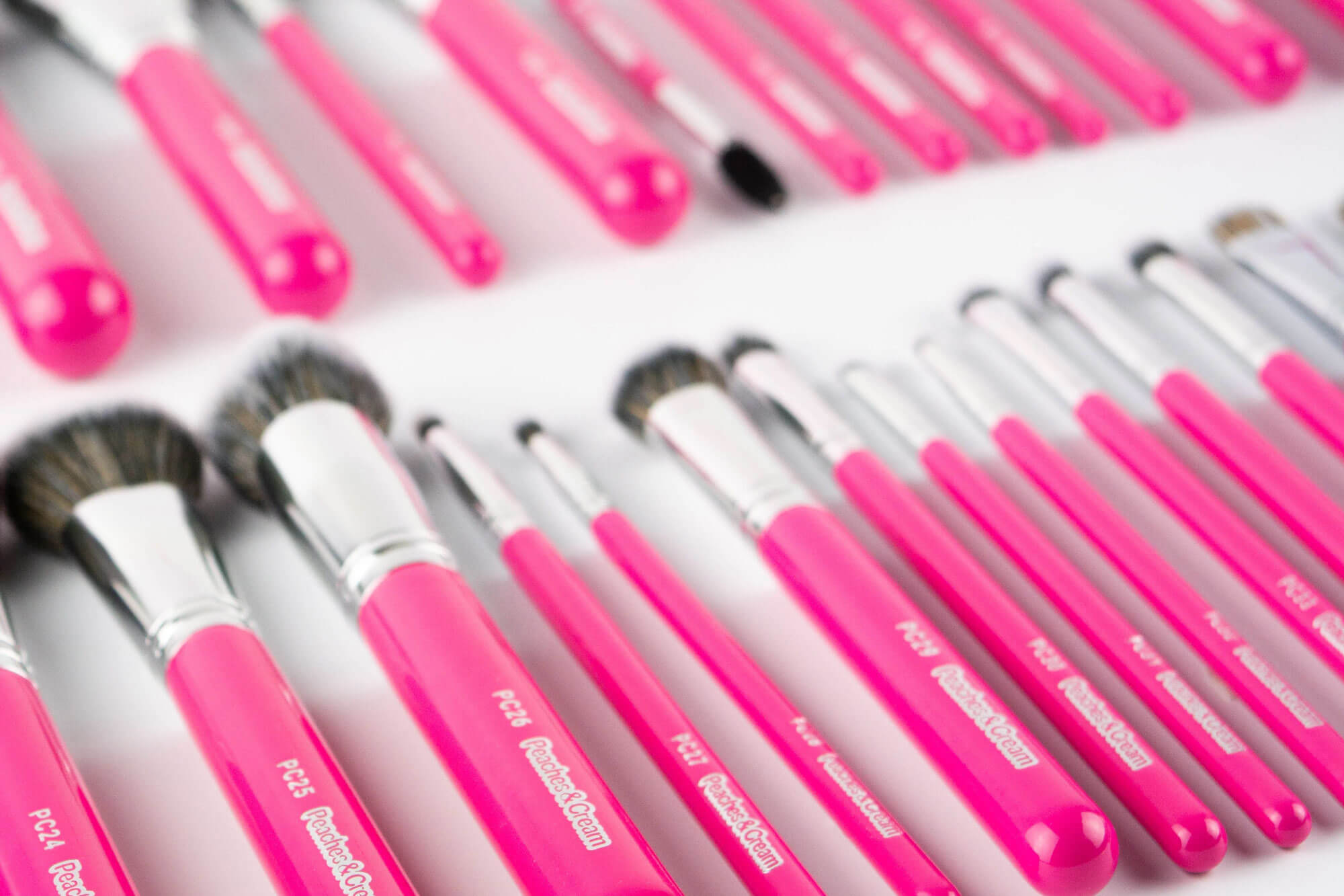 Various pink-handled makeup brushes with silver ferrules and purple bristles are arranged in a scattered pattern on a bright blue background.