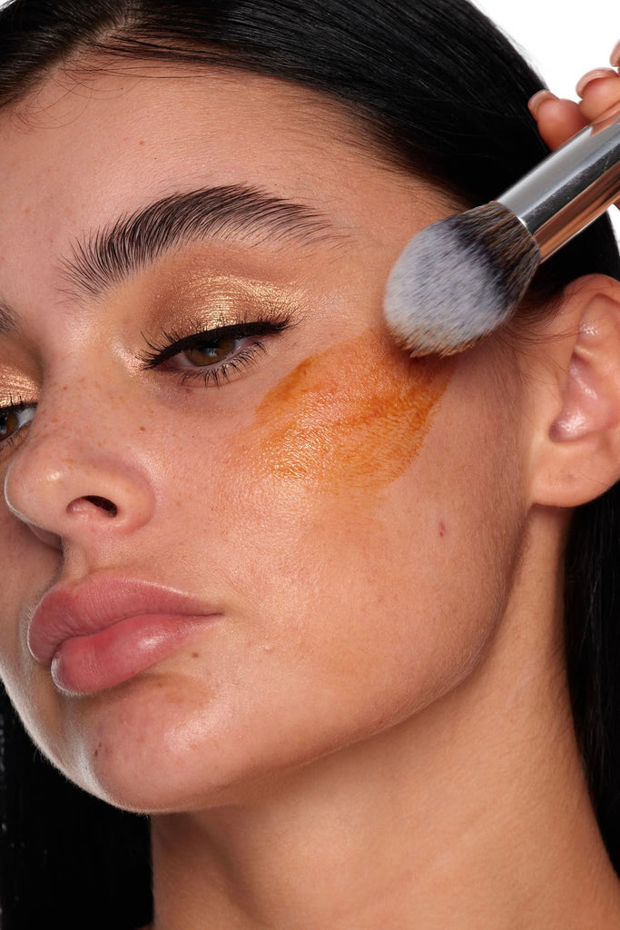 A woman uses a makeup brush to apply Peaches and Cream’s Sun Tint bronzer to her cheek, achieving a sun-kissed look. Her smooth skin, defined brows, and shimmery gold eyeshadow contribute to an overall natural, glowing appearance.