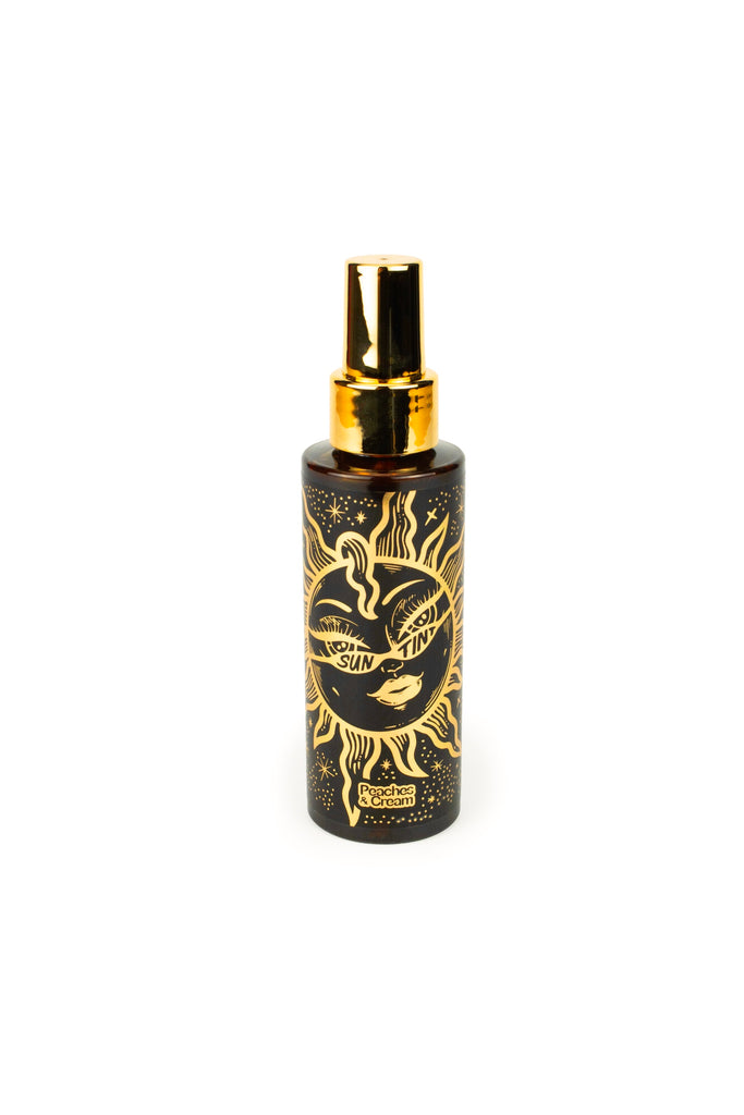 A spray bottle in black and gold from Peaches and Cream, labeled Sun Tint, provides a sun-kissed appearance. Decorated with celestial suns and stars, it boasts a shiny gold cap, making it the perfect bronzing gel for a radiant glow.