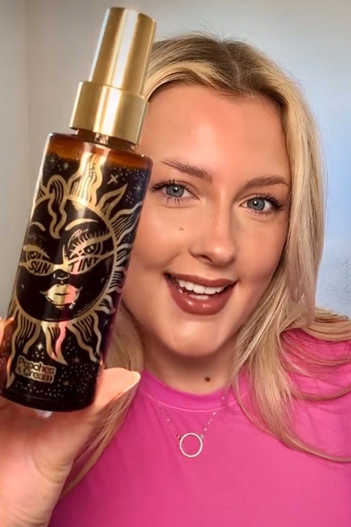 A person with long blonde hair and a bright smile holds up a decorative bottle of Sun Tint by Peaches and Cream, featuring a celestial-themed design. They're wearing a pink top, circular pendant necklace, and have achieved the perfect sun-kissed look while posing in a well-lit indoor setting.